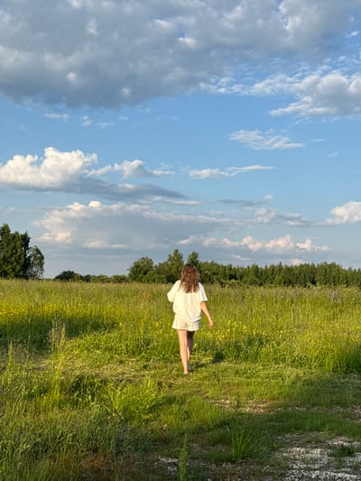 A young woman with curly brown hair, wearing a white t-shirt and white shorts, walks away from the camera through a tall, green grassy field. The field is dotted with small yellow wildflowers. In the background, a dense line of green trees forms the horizon. Above the trees, a bright blue sky is filled with fluffy white and gray clouds. The scene appears to be set in a rural area, possibly near the village of Koloksha, Russia, given the natural landscape. The lighting suggests it is late afternoon or early evening, with the sun casting long shadows on the grass.