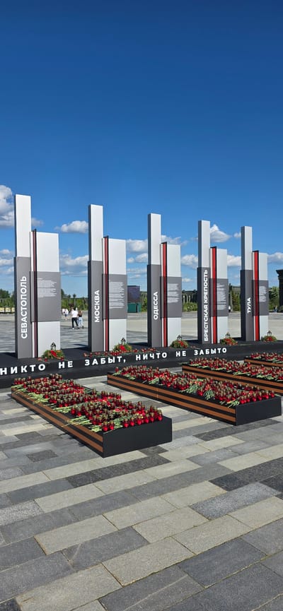 The image depicts a memorial site with a row of tall, modern obelisks against a bright blue sky with scattered clouds. Each obelisk has vertical bands of white and gray, with a thin red stripe. On the front of each obelisk, in white lettering, are names of cities: "СЕВАСТОПОЛЬ", "МОСКВА", "ОДЕССА", "БРЕСТСКАЯ КРЕПОСТЬ", and "ТУЛА". Below these obelisks, a black banner displays white text that reads "НИКТО НЕ ЗАБЫТ, НИЧТО НЕ ЗАБЫТО" (Nobody is forgotten, nothing is forgotten). In front of the banner are several long, black rectangular planters filled with numerous red candles and red carnations, arranged in rows. The planters are lined with orange stripes. The ground is paved with dark gray and lighter gray rectangular stones laid in a pattern. In the background, there are some green trees and other structures, including what appears to be a distant building with arched windows. A few people are visible walking in the middle ground, some facing away from the camera. The overall scene suggests a solemn commemoration of historical events or fallen heroes. The location is identified as being in the city of Zelenaya Roshcha, Russia.