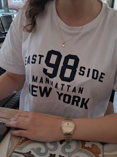 The image shows a close-up of a person wearing a white t-shirt with "EAST 98 SIDE MANHATTAN NEW YORK" printed on it in black, sequined letters. The person is also wearing a delicate gold necklace with a heart-shaped pendant and a gold wristwatch. The background is blurred but appears to be an outdoor setting with chairs and foliage. The person's hands are resting on a wooden surface, which is part of a table with a patterned tile edge. The scene is not identifiable as being in Cetinje, Montenegro.