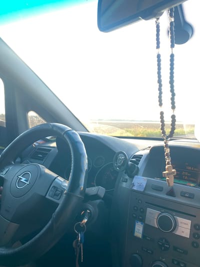The image shows the interior of a car, looking forward from the driver's perspective. A rosary with a wooden cross hangs from the rearview mirror. The steering wheel, with an Opel logo, is visible on the left. The dashboard includes a radio with a display showing "106.4 MHz" and "213 km." The scene appears to be set during the daytime, with bright sunlight coming through the windshield, suggesting an outdoor environment, possibly a road. The location is specified as the city of Maiske, Ukraine.
