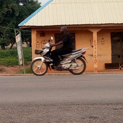 The image depicts a man riding a silver and blue motorcycle along a paved road in Mararaba, Nigeria. He is seen in profile, wearing dark clothing, and appears to be in motion, driving past a building on the right. The motorcycle has a small basket mounted on its front.

In the background, a single-story building with ochre-colored walls and a light-colored corrugated metal roof with a blue trim is visible. It features a dark doorway on the right. On the building's wall, painted vertically in orange letters, is the text "KO+700". Below the roofline and above the doorway, there are crude painted outlines of objects, resembling gas cylinders or lanterns. Next to one of these outlines, some indistinct text, possibly "(CJ)", can be seen.

To the left of the building, a green-framed sign with a white background stands by the roadside, displaying black text that is largely unreadable. Beyond the building and sign, lush green trees and foliage are visible. The road in the foreground is paved and appears dry.

The scene is lit by natural daylight, suggesting it is daytime, possibly morning or afternoon. The weather appears to be bright but diffused, with no strong direct shadows. The overall atmosphere conveys a moment of daily activity in a semi-urban or rural setting.