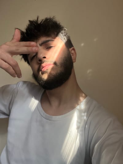 The image displays a single male individual positioned indoors in Joubar Municipality, Syria. The subject is a young adult with dark hair and a dark beard, wearing a light-colored, long-sleeved crew-neck shirt. His left hand is raised, with fingers resting above his left eyebrow. Direct natural sunlight, originating from the upper left, casts a prominent diagonal stripe of illumination across the right side of his face and upper body, creating distinct highlights and shadows. The background is a plain, light-colored wall, with subtle, irregular light patterns visible on the right side. The individual maintains a neutral facial expression, looking towards the viewer.