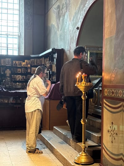 In the interior of a religious building in Veliky Novgorod, Russia, a man with long, gray hair and glasses, wearing a light-colored checkered shirt and light-colored pants, stands facing left, his right hand raised. He is positioned in front of a dark shelf filled with numerous religious icons. To his right, another man in a dark jacket and dark pants stands facing forward, his head turned to the left, obscuring his face. He is looking towards an ornate golden candelabrum with lit candles. A woman, mostly obscured, stands behind and to the right of the man in the dark jacket. The environment includes a large window on the left with a grid pattern, a section of a fresco on the wall above, and decorative elements on a curtain to the right. The floor is tiled.
