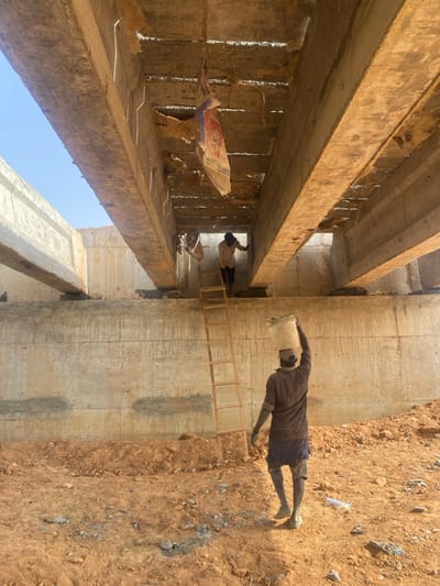 An image shows a construction site with two men. One man stands on the ground, adjacent to a concrete structure, with a ladder placed against it. He wears dark clothing and a cap. The other man is on a ladder inside the concrete structure. The structure appears to be a bridge under construction, composed of concrete beams. Debris hangs from some of the beams. The ground outside is dirt with scattered litter.