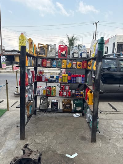 The image depicts an outdoor retail display of automotive products situated on a concrete surface in Badore, Nigeria. A prominent four-tiered, black metal shelving unit, positioned centrally, holds numerous plastic containers of engine oil, transmission fluid, and other automotive lubricants in various colors (yellow, red, silver, black, blue, green). Brands such as Mobil Super are visible on some containers. Cans of spray products and smaller metal cans are also arranged on the lower shelves.
In the immediate foreground to the lower left, a discarded engine block or a similar heavy metallic automotive component rests on the textured concrete ground, which shows minor debris including small pieces of white paper.
To the left of the display, a paved street is visible, bordered by a white line. A faded "NO PARKING" sign is attached to a pole, and another sign, partially obscured, indicates "UTME & WAEC". A stack of black tires is visible behind these signs, adjacent to a low-rise building with a brown roof. A metal stanchion with a blue rope barrier is present near the street.
In the background to the right, a dark-colored SUV, likely a Subaru Forester model, is parked behind the shelving unit, with its left side and roof rack visible. Beyond the vehicle, a light-colored building with a green-trimmed roof is present. Overhead utility lines and poles crisscross against a partly cloudy, light blue sky. A palm tree is visible behind the shelving unit in the mid-distance. The scene is devoid of any visible people or ongoing activity.