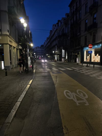 The image depicts a street scene in Brussels, Belgium, at dusk or night. The sky is a deep, clear blue, indicating low light conditions. The street is paved, with a cobblestone sidewalk on the left and asphalt on the right. A yellow-painted bike lane with a white bicycle symbol is prominently visible on the right side of the road. Streetlights illuminate the scene, casting a warm glow on the buildings lining the street. The buildings are multi-story, with traditional European architecture, featuring balconies and large windows. Some shop windows are lit, displaying merchandise. A few cars are present on the road, with headlights on. Two young men are standing on the sidewalk on the left, appearing to be engaged in conversation. There is a traffic sign on the right side of the street, a red circle with a horizontal white bar, indicating a prohibition, likely a "no entry" sign. The overall impression is that of a quiet urban street in the evening, possibly after business hours, with some activity on the sidewalk and street.