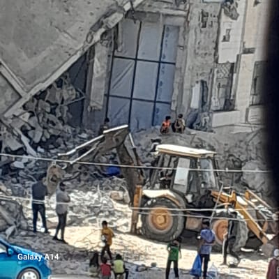 The image depicts a scene in Khan Yunis, Palestinian Territories, amidst significant destruction. A partially collapsed building stands adjacent to a pile of rubble. A yellow construction vehicle, possibly a bulldozer or backhoe, is present near the debris. Several people are visible, some appearing to be amidst the rubble.