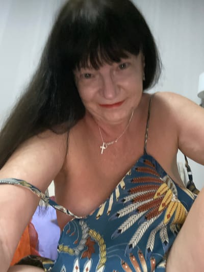 A close-up shot shows a woman with long, dark hair and bangs. She is wearing a dress with a colorful pattern of feathers. A silver cross necklace hangs around her neck. The background is blurred and appears to be an indoor setting. The image is undated and unlocated.