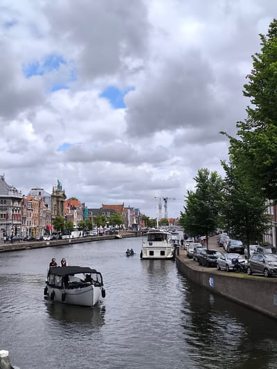 This is a medium shot, taken from a slightly elevated perspective, of a canal in Haarlem, Netherlands. On the left bank of the canal, a row of old, multi-story buildings with ornate facades line the water. Several people are walking on the sidewalk in front of these buildings. A small tour boat with a black canopy is moving down the canal, carrying at least three people. On the right bank, a tree-lined promenade runs parallel to the canal, with a street behind it. Cars are parked along this street. In the background, several cranes are visible, suggesting ongoing construction or development. The sky is overcast with dramatic clouds.