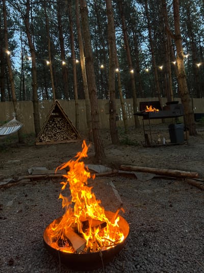 The image depicts an outdoor scene in a wooded area. In the foreground, a fire burns in a circular metal fire pit filled with wood. The ground is covered in gravel and scattered pinecones. In the mid-ground, a triangular structure of stacked firewood stands to the left, with a hammock hanging nearby. To the right, a barbecue grill with a fire inside is set up on legs, accompanied by a trash can and some spray cans. String lights are strung between trees in the background, illuminating the dusk sky. The location is identified as Ivachiv Horishnii, Ukraine. No people are visible in the image.