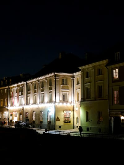 The image shows a street scene at night in Warsaw, Poland. The buildings are lit up, and there are a few people walking on the sidewalk. In the foreground, there is a dark area that appears to be a river or canal. A dark-colored car is parked on the street in front of one of the buildings. The building on the right has a sign that reads "BARSZCZ & BORSZCZ". The architectural style of the buildings suggests they are historical, possibly part of the Old Town.
