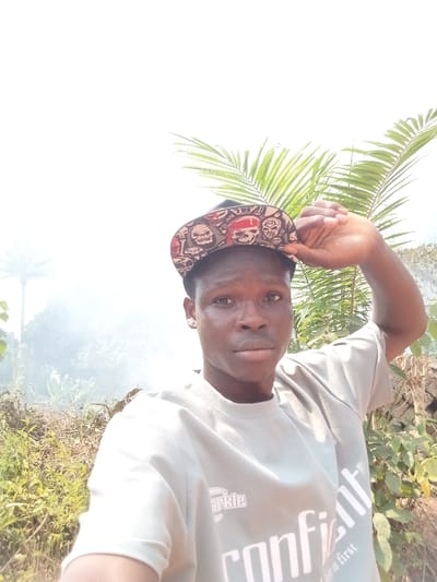 A young adult male, of dark complexion and short dark hair, is depicted in a close-up, chest-up outdoor shot in Eziudo, Nigeria. He wears a light grey crew-neck t-shirt with partial text "Sparkle" and "confident" visible, and a black baseball cap patterned with multiple stylized skulls, some featuring red bandanas, worn backward. His right hand is raised, gripping the brim of the cap on the right side of his head. He gazes directly forward with a neutral expression. The background consists of an overexposed bright white sky, dense green vegetation, including prominent palm fronds behind his head, and the silhouette of a distant palm tree on the far left. A distinct plume of white smoke or haze is visible in the mid-ground to the left of the subject, partially obscuring vegetation. The immediate ground appears to be an unpaved area with sparse dry and green plant growth.