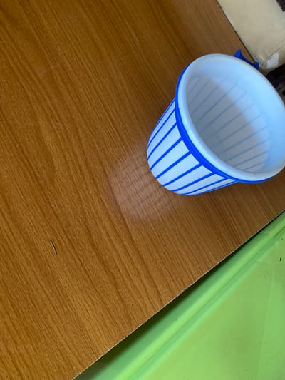 A white plastic cup with blue stripes is placed on a wooden surface. The cup is oriented towards the right side of the frame and appears to be empty. The wooden surface has a visible wood grain pattern. In the background, to the right of the cup, a portion of a light-colored fabric object is visible, possibly furniture. Below the wooden surface, a bright green surface is partially visible. There are no people or discernible activities taking place. The scene depicts an indoor setting with everyday objects. The image location is Jauro Yinu, Nigeria.