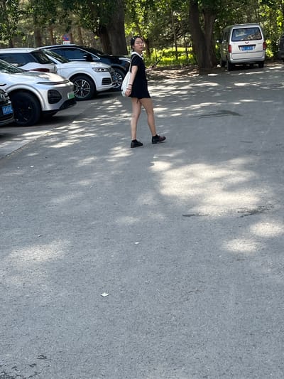 A woman in a black dress stands in the street next to a line of parked cars in Changchun City, China. A silver vehicle with license plate "吉A·18537" is visible.