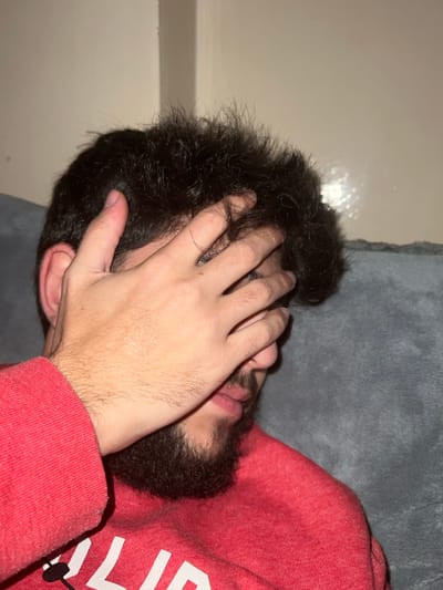 A person with dark hair and a beard lies on a gray surface, possibly a couch or bed. The person's face is obscured by their hand, palm facing the camera. They wear a red sweatshirt, the fabric slightly rumpled. The background is a pale, out-of-focus wall with a faint light reflection. The image is cropped closely, focusing on the person's head and upper body.