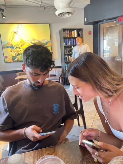 The image shows three individuals inside a commercial establishment, likely a cafe or coffee shop. In the foreground, a young adult male with dark curly hair and a short beard is seated at a wooden table, wearing a brown t-shirt with "Stereo" and Korean characters ("사운드") printed in blue. He is looking down at a light blue smartphone held in his right hand. To his right, a young adult female with long light brown hair is seated, wearing a white top and multiple silver necklaces. She is holding a light-colored smartphone and looking towards the male's general direction or his phone. Both foreground subjects are focused on their mobile devices. In the background, an older adult male with gray hair and a beard, wearing a light blue collared shirt, stands behind a dark shelving unit filled with various mugs and tumblers. He is looking to the right. On the left background wall, a large, abstract painting depicting a yellow sun-like form over a colorful geometric landscape is visible. A white ceiling fan with a light is mounted overhead. A wooden door with a glass panel and a partial "EXIT" sign is visible on the far right. The specific location within the city of Washington, United States, cannot be determined from the image content.