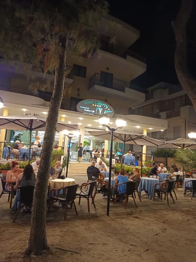 This image depicts an outdoor dining area at the Hotel Liberty in Roseto degli Abruzzi, Italy, at night. The hotel features a prominent illuminated sign that reads "Hotel Liberty" along with "Restaurant" and "Fashion Cafe." People are seated at various tables covered with blue tablecloths, enjoying their meals and drinks under the glow of overhead lights and street lamps. Some tables are partially obscured by trees and umbrellas, creating a cozy and somewhat dimly lit ambiance. A few individuals are visible standing and walking within the restaurant's vicinity, contributing to the lively atmosphere of an evening at a popular establishment. The ground appears to be sandy or dirt, suggesting the location might be near a beach or a similar natural setting.