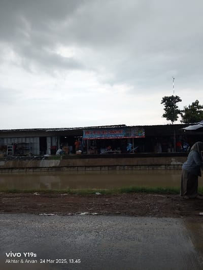 The image shows a view of a street in Kecamatan Haurgeulis, Indonesia (latitude: -6.419, longitude: 107.972). There are multiple shops or stalls lining the street. A sign above the shops reads "Baju & Tas Perempuan", which means "Women's Clothes & Bags" in Indonesian. The street is wet and muddy with a canal running along the side. A few people can be seen walking in the distance. The sky is overcast with grey clouds. The time is 13:45.