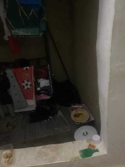 The image depicts a dimly lit interior space, possibly a room or storage area, in Howayi, Nigeria. The scene is cluttered with various items, suggesting a state of disarray or storage. In the foreground, a beige-colored surface is visible, with a small white and brown object on it, potentially a food item. To the left, a white, circular object with a dark "U" shape is present, possibly a lid or base. Next to it, a small white cup is standing upright. A green object resembling a comb lies on the surface. A small box with a yellow and red design is also visible. In the background, a dark backpack is placed on the floor. Various pieces of clothing are scattered around, including a red garment with white soccer ball designs, a gray garment with black patterns, and other folded clothes. A metal shelf unit is partially visible, supporting some of these items. The walls appear to be a light, textured material. The overall impression is one of a utilitarian and somewhat neglected space.