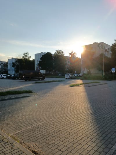 The image depicts a street scene in Baranavichy, Belarus, likely in a residential area. A paved parking lot occupies the foreground, made of rectangular interlocking bricks. Several parked vehicles are visible, including a dark-colored panel van, a white van, and several passenger cars. In the background, apartment buildings of various heights are visible, some with visible architectural details and murals. Trees and greenery are interspersed between the buildings and around the parking area. The sun is low in the sky, casting long shadows and creating a warm, golden light that flares across the image, particularly over the right side of the scene. A blue parking sign with a white "P" and a bicycle symbol is visible on a pole on the right side of the image. The sky is a clear blue with a few wispy clouds. The overall impression is a quiet, late afternoon or early evening urban setting.