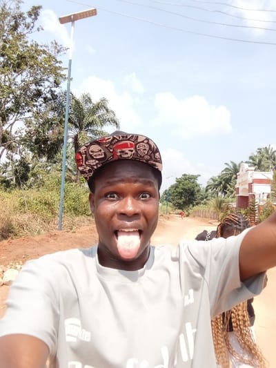 A forensic description of the image, set in Eziudo, Nigeria, reveals a daytime outdoor scene. The primary subject is a dark-skinned male positioned in the foreground, engaged in taking a selfie. He wears a light grey short-sleeved t-shirt and a multi-colored snapback baseball cap adorned with skull motifs. His eyes are wide, and his tongue is extended.
To the right and slightly behind the main subject, the back of another individual is partially visible, characterized by long, light-colored braided hair and a dark garment. Further down a reddish-brown dirt road, a small, blurry figure in red clothing is discernible in the distance.
The environment features a clear blue sky with scattered white clouds. On the left side, a tall, slender pole supports a solar-powered street light. Dense green foliage, including palm trees and other deciduous trees, lines both sides of the dirt road. In the mid-ground on the right, a distinctive white and reddish-brown architectural structure, possibly an ornate gate or entrance, stands beside the road.