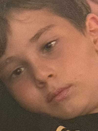 A close-up, diagonally oriented image captures the face of a single individual, appearing to be a juvenile or young person, within an indeterminate indoor setting in Veintisiete de Abril, Costa Rica. The subject has fair to light-brown skin, short-cropped dark hair visible on the top and right side of the head, and dark eyes. The left eye, positioned toward the center of the frame, shows a distinct light reflection. The right eye is partially visible towards the upper left. Dark eyebrows are present above both eyes. The nose and mouth are visible in the lower half of the face, with lips closed in a relaxed posture. A dark-colored garment is visible at the bottom and lower-left, featuring a partial light-colored, possibly gold or yellow, graphic detail on the lower right side of the frame. The background is uniformly dark and out of focus. No specific actions or interactions are discernible beyond the subject's gaze.