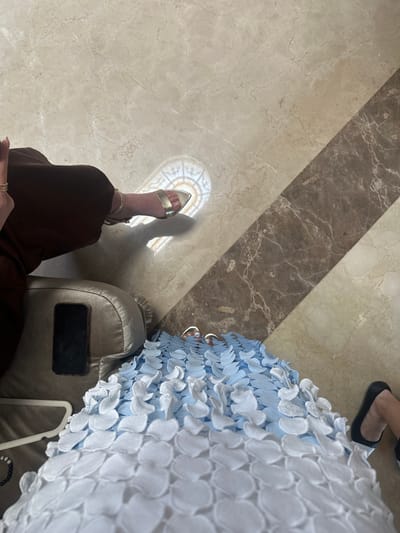 The image depicts an indoor scene in Monterrey, Mexico, focusing on the lower bodies of two individuals and their immediate surroundings. The floor is composed of polished marble, with a predominant light beige section showing subtle veining, diagonally bisected by a strip of darker, mottled brown marble. A distinct circular, ornate sunlight reflection, resembling a decorative window pattern, is cast onto the lighter marble near the center-left. A sharp diagonal shadow line extends across the floor from the upper right.
On the left, a person is seated, wearing dark brown wide-leg trousers and a single gold metallic open-toed sandal on their right foot. A hand with multiple thin gold rings is partially visible above the leg. Adjacent to this individual is a beige upholstered armrest, upon which a black smartphone rests. A white plastic garment hanger and a black beaded bracelet are present on the floor to the left of the armrest.
To the right, the lower portion of a second person's attire is visible. This person wears a light blue and white garment featuring numerous layered, petal-like appliques. Their feet are partially visible, wearing metallic open-toed sandals. A black flat shoe is situated on the floor near their right foot.