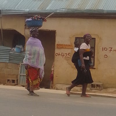 In Gombe, Nigeria, two women are seen walking on a paved road in front of a building with a corrugated metal roof and a pale yellow, textured wall. The woman on the left is older and carries a blue bowl filled with items on her head, balanced with a stick. She wears a purple scarf draped over her shoulders and a colorful red and yellow patterned skirt. She is smiling. The woman on the right is younger and carries a baby on her back. She is wearing a pink headscarf, a white t-shirt with a yellow detail, and a long dark skirt. She is also smiling.
The scene appears to be during the daytime. There are some notable details on the wall of the building. A wooden sign with black lettering reads "AZOA SAYA- RUWA 1". Below this, also written in dark paint on the wall, is "C+475". Further to the right, "10.7m" is written in red, and a circular symbol is present. The building's entrance is visible, along with some objects to the left, including a blue bucket and stacked materials. In the foreground, a concrete curb separates the road from the area in front of the building. The overall impression is a snapshot of daily life in a Nigerian town.