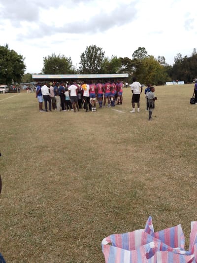A rugby team in maroon and blue uniforms is gathered around a group of people, likely fans and staff, on a grassy field under an overcast sky. A few individuals in casual attire stand apart from the main group. A small child walks toward the huddle. A covered seating area can be seen in the background, indicating a sports ground. A striped bag sits in the foreground, partially obscuring the view.