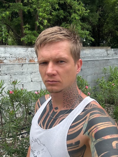 A man with blond hair and a light complexion is taking a selfie outdoors. He is wearing a white tank top and a silver chain necklace. His neck and arms are covered in intricate black tattoos. In the background, there is a white brick wall and lush green foliage with small pink flowers. The man appears to be standing on a path covered with white stones. The scene is likely located in Ban Tong Kai, Thailand.