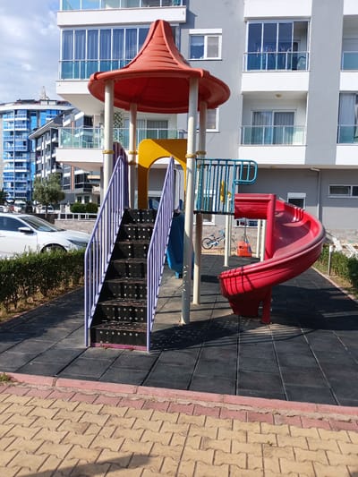 The image depicts an outdoor playground with a red spiral slide, a purple staircase, and a yellow slide, all part of a larger play structure. The playground is situated on a black rubberized surface, with a border of interlocking beige and pink pavers in the foreground. Behind the playground, a modern grey building with balconies and large windows rises. To the left, a blue building with many windows and a distinctive curved design is visible, along with parked white cars and a dense green hedge. The sky is blue with some white clouds, suggesting a clear, sunny day. The lighting indicates it's daytime, likely late morning or early afternoon. There are no people visible in the image, and no text is present. The scene appears to be a residential area, possibly an apartment complex, in Alanya, Turkey, given the architectural styles and the presence of a playground.