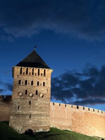 The image shows a tall, brick tower and a section of the wall of the Veliky Novgorod Kremlin, also known as the Detinets, at dusk. The sky is a deep blue with dark, cloudy formations. The tower, constructed of red brick, has a dark, conical roof topped with a weather vane. It features numerous small, arched windows and some decorative circular elements on its facade. The wall, also made of brick, extends horizontally to the right of the tower and is illuminated by artificial lights, highlighting its texture and battlements. A grassy slope leads up to the base of the wall and tower. No people are visible in the image. The scene is a nighttime view of a historic fortress in Veliky Novgorod, Russia.