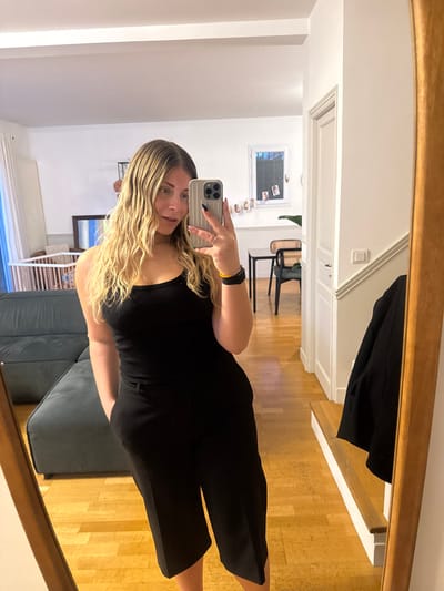 An indoor mirror selfie depicts a blonde female, approximately 20-30 years old, standing in a residence in Rueil-Malmaison, France. She is wearing a black ribbed tank top and black cropped wide-leg trousers. Her right hand, with dark nail polish, holds a light gray smartphone featuring a ridged case and triple-lens camera, used to capture the image. Her left hand is placed in her trouser pocket, and a yellow band is visible on her left wrist. She stands on a light-colored wooden parquet floor, reflected in a full-length mirror with a light wooden frame. The background, visible in the mirror's reflection, includes white walls, a partial view of a dark gray fabric sofa, a wooden baby crib, a dark-framed mirror, a white doorway, a dark dining table with two dark chairs, and a window with hanging photographs and a potted plant.
