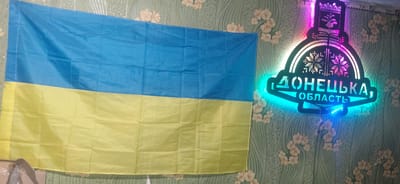 A Ukrainian flag hangs on a wall with a floral patterned wallpaper in Ketrysanivka, Ukraine. To the right of the flag is a neon sign with the text "ДОНЕЦЬКА, ОБЛАСТЬ" in Cyrillic letters, which translates to "Donetsk Oblast". The sign is illuminated with blue and pink neon lights. The flag is partially visible, with its blue top half and yellow bottom half clearly displayed. The wallpaper has a light green background with light pink floral designs. The image captures a still scene, with no people or apparent actions occurring.