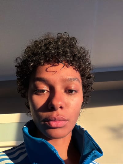 A close-up, head-and-shoulders selfie shows a young woman with curly, dark hair, looking directly at the camera. She is wearing a blue jacket with white stripes. The lighting suggests it is late afternoon or early morning, with warm sunlight casting shadows on her face and the wall behind her. The background appears to be an indoor setting with a wall and a window. There are no specific landmarks visible that would indicate the location within Champs-sur-Marne, France.