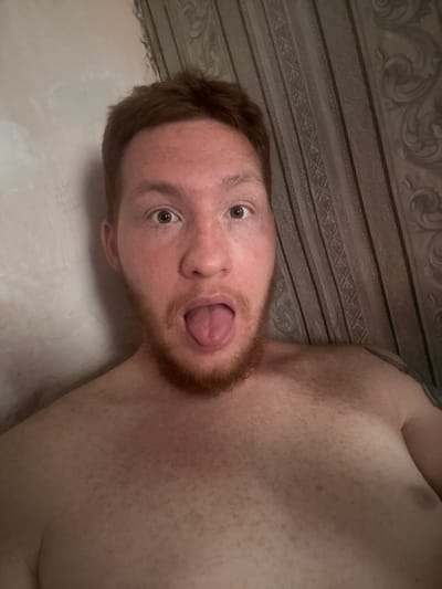 A close-up, selfie-style photograph shows a young, pale-skinned man with red hair and a light beard sticking his tongue out. He is shirtless and appears to be indoors, leaning against a wall with textured wallpaper on the right side of the frame. The lighting is dim and casts shadows, obscuring some details. There is no identifiable information to indicate the location as Makiivka, Ukraine.