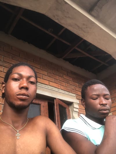 Two dark-skinned men are positioned in the frame. One wears a light-colored, collared shirt with white stripes, eyes closed. The second is shirtless, wearing a gold chain with a cross pendant. They appear to be outside. A brick building and visible ceiling joists form the backdrop. The camera angle is low, tilted upward.