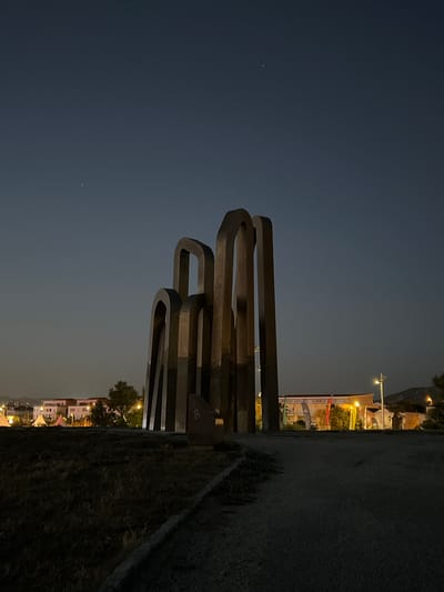 The image depicts a modern sculpture at night, situated in a park-like setting. The sculpture, made of dark, curved metal panels, dominates the center of the frame. Behind the sculpture, a path leads towards buildings that are illuminated with artificial lights. Trees are visible between the sculpture and the buildings. The sky is dark, with a few faint stars visible, suggesting a clear night. The ground in the foreground is covered with dry grass and a gravel path. The overall atmosphere is quiet and serene.