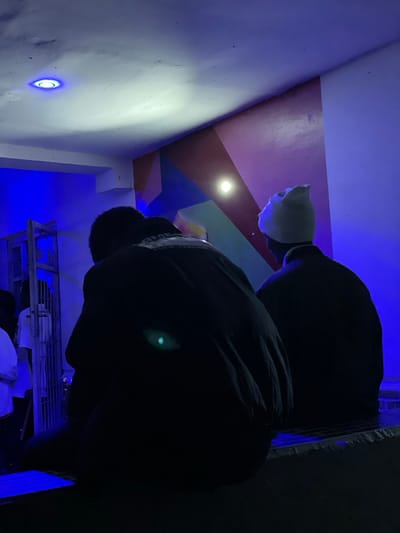 The image depicts an indoor scene with low lighting, dominated by blue and purple hues. Two individuals are seated, viewed from behind. The person on the right wears a light-colored beanie and a dark jacket. The person on the left wears a dark jacket with a visible white emblem on the back. In the background, a metal gate or fence with intricate designs is visible, illuminated by blue light. The wall behind the individuals features abstract geometric shapes in shades of red, purple, and blue. A circular light fixture with a bright blue glow is present on the ceiling. The overall environment suggests an urban setting within the city of Durimi, Nigeria, potentially a public or private gathering space, illuminated by artificial lighting.