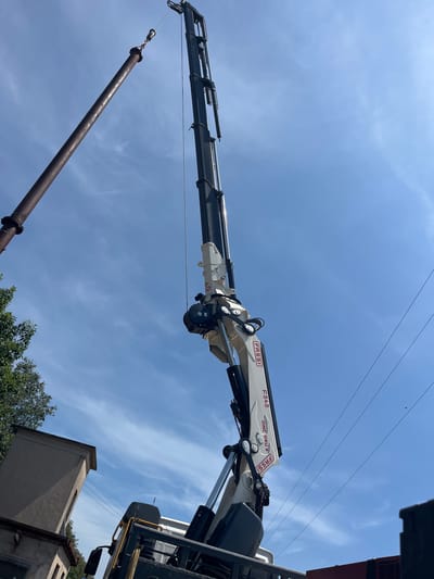 In Castiglione delle Stiviere, Italy, a large industrial pipe, rusted and weathered, is being lifted into the air by a white and grey Fassi F545 OMC GRU crane mounted on a truck. The crane's hydraulic arm is extended upwards, suspending the pipe via a cable and attachment system. The sky is a clear blue with scattered white clouds. In the lower left of the image, the top of a tan-colored building is visible, partially obscured by green foliage from a tree. Power lines stretch across the sky in the background. The scene depicts industrial work taking place outdoors.