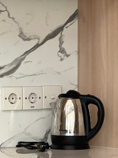 A close-up, eye-level shot features a chrome and black electric kettle with a visible brand name "Greflide" on its side, resting on a white surface. To the left of the kettle are three white electrical outlets mounted on a marbled white backsplash with gray veining. A black electrical cord with a plug lies on the white surface in front of the outlets. The background to the right of the backsplash is a light brown wooden panel. The image appears to be taken indoors, possibly in a kitchen or common area, with no people present. The overall impression is one of a functional domestic setting.