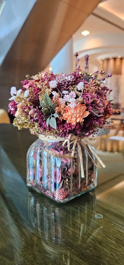This is a close-up, top-down shot of a dried flower arrangement placed next to a glass jar filled with potpourri. The flower arrangement is comprised of various dried flowers and foliage in shades of deep red, burgundy, pink, and gold, with a single larger, orange-hued flower as a focal point. A thin, straw-like ribbon is tied around the base of the bouquet, securing it to the side of the glass jar.
The glass jar is hexagonal and appears to be made of clear, ribbed glass. It is filled with a mixture of dried plant material, likely potpourri, in various shades of pink, red, and brown, with some darker, almost black pieces. The contents of the jar are layered, suggesting it might be a decorative item for scent.
The items are placed on a reflective, dark surface that appears to be glass or polished wood, showing faint reflections of the objects and the environment. In the background, out of focus, are soft, warm lights, suggesting an indoor setting, possibly a hotel lobby or a well-lit interior space. The lighting creates a gentle, ambient glow. The overall impression is one of elegant and natural decoration. There is no visible text in the image. The context of Doha, Qatar, suggests this might be an interior decor element in a public space or private residence within the region.