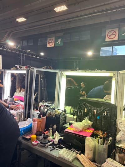 The image depicts an indoor preparation area, likely a backstage or dressing room setup, located in Issy-les-Moulineaux, France. The scene features multiple vanity stations with illuminated mirrors.

In the foreground, a large table functions as a shared makeup and styling workstation. It is covered with numerous cosmetic products, including bottles of lotions and cleansers (one labeled "NUXE"), various makeup palettes (one prominent pink palette possibly labeled "Waterproof"), tubes, compacts, and brushes stored in several clear and black organizers. Cotton swabs are contained in a clear plastic box, and a blue and white tissue box is present. A dark-clad arm and partial torso of an individual are visible on the far left, reaching towards items on the table.

Three lighted vanity mirrors are visible, showing reflections of individuals. In the leftmost mirror, a person with dark hair is reflected, appearing to use a laptop with a pink casing. In the rightmost mirror, two individuals are reflected: one person with curly reddish-brown hair wearing a black shirt with "TEAM" written on the back, and another individual, likely a client or model, seated and leaning forward, wearing a blue top. This suggests makeup application or hairstyling is in progress.

The environment features dark drapes or walls in the background, with metal trussing visible horizontally. The ceiling is industrial, with exposed structures and rectangular fluorescent light fixtures. Hanging from the ceiling are multiple square signs: a "No Smoking" symbol (red circle with a crossed-out cigarette) and "Emergency Exit" symbols (green square with a running person). Windows or skylights in the upper right reveal a glimpse of an exterior building structure or sky.