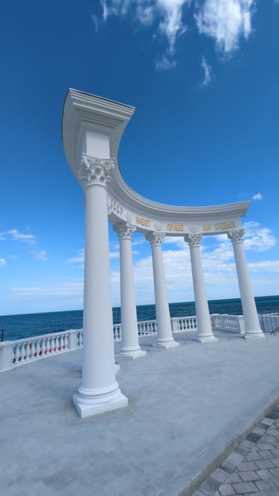The image depicts the Arch of the Soviet Union, a white neoclassical structure located in Alushta, Crimea, Ukraine. The arch is adorned with Corinthian columns and inscribed with the phrase "СССР ИМЕЮТ ПРАВО НА ОТДЫХ" (The USSR has the right to rest) in gold lettering. The structure overlooks the Black Sea, with blue water and a bright sky with scattered clouds visible in the background. The foreground shows a paved area with a white railing. There are no people visible in the image.