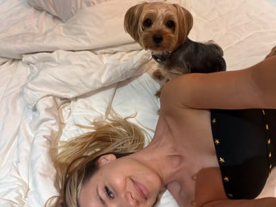 The image shows a woman and a small dog lying on a white bed. The woman is lying on her side, with her head resting on the pillow and her body angled towards the camera. She has blonde hair and is wearing a black bikini top with gold stars. Her face is visible, and she appears to be smiling. The dog is a Yorkshire Terrier and is sitting up on the bed next to the woman, looking at the camera. The setting appears to be a bedroom with white linens. The lighting suggests it might be daytime, possibly indoors. There is no visible text in the image.