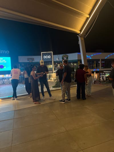 This image depicts a group of people standing on a tiled walkway at night, likely in an urban or commercial setting. The scene is illuminated by overhead lighting and the glow from building facades. The people appear to be casually dressed and are engaged in conversation or looking at their phones.
In the background, there are storefronts with illuminated signage, suggesting a shopping mall or entertainment complex. One sign clearly reads "OCIO," which translates to "Leisure" in Spanish. Another sign features the word "BOWLING" with a cartoonish graphic, indicating a bowling alley. The overall atmosphere appears relaxed and social. The time of day is nighttime, as indicated by the dark sky and artificial lighting.
The people in the foreground are diverse, with individuals of various ages and genders. Their clothing is casual, consisting of t-shirts, jeans, and dresses. Some are holding cell phones, possibly communicating or browsing. The setting appears to be outdoors, possibly a plaza or pedestrian area connected to shops and entertainment venues. The ground is paved with large, square tiles. The lighting casts soft shadows, contributing to the nighttime ambiance. The presence of "OCIO" and "BOWLING" signs strongly suggests that the location is an entertainment or leisure complex in Alcorcón, Spain.