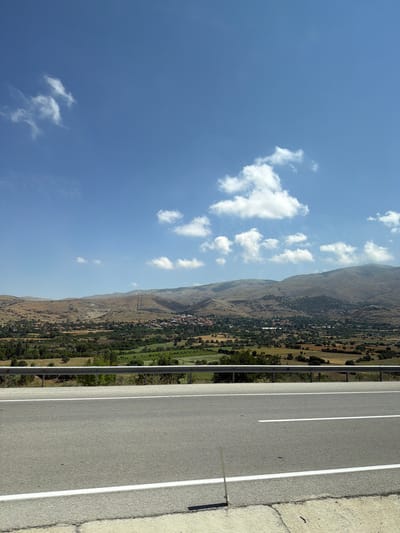 The image depicts a wide shot of a rural landscape under a bright blue sky with scattered white clouds. In the foreground, a paved road with white lane markings stretches horizontally across the frame. A metal guardrail separates the road from the embankment. Beyond the road, the landscape unfolds into rolling hills covered with dry, scrubby vegetation and patches of green fields. A small village, characterized by numerous red-tiled roofs, is nestled in the valley below, surrounded by trees. In the background, larger, more rugged mountains form a backdrop against the sky. The scene appears to be taken from an elevated vantage point, likely from a vehicle traveling on the road. The environment suggests a warm, sunny day in a semi-arid region. There are no people visible in the image. The location is identified as being in the vicinity of Çavdır, Turkey.