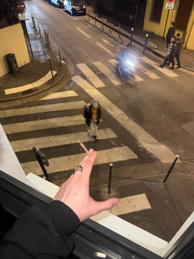 The image depicts an elevated view of a wet urban street at night in Asnières-sur-Seine, France. In the immediate foreground, a light-skinned left hand, wearing a metallic ring on the ring finger, extends over a dark window frame and concrete sill, holding a lit, hand-rolled cigarette between the thumb and index finger. A discarded cigarette butt is visible on the sill.
The street below features a prominent pedestrian zebra crossing with white stripes. The asphalt pavement appears reflective, indicating wet conditions. Parked on the left side of the street are three dark-colored vehicles. A scooter or motorcycle with bright headlights is in motion, traversing the pedestrian crossing from left to right, exhibiting motion blur.
Several individuals are present on the street level. One person, blurred by movement, is walking across the pedestrian crossing, wearing a dark jacket, a light-colored top, and a light hat. Further down the street on the right, two other individuals are standing on the sidewalk adjacent to a yellow building.
Street furniture includes multiple dark bollards with light tops lining the sidewalks. On the left, a sign with a red circle, diagonal bar, and bicycle symbol is partially visible, displaying the word "SAUF." Another square sign on the right also includes "SAUF 8H-18H" and a bicycle symbol. A beige-colored building with a curved corner and a recessed archway is on the left, while a yellow building with dark-framed windows stands on the right. A green public waste bin is located near the beige building's corner. Artificial street lighting illuminates the scene.