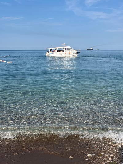 The image depicts a clear, blue sky over a tranquil sea with a pebble beach in the foreground. A white passenger boat carrying several individuals is navigating the water in the mid-ground. Further in the distance, two cargo ships are visible on the horizon. The water is exceptionally clear, allowing the submerged pebbles to be seen distinctly. This scene is consistent with the coastline of Bar, Montenegro, known for its clear waters and pebble beaches.