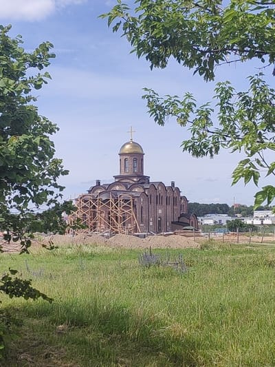 The image depicts an unfinished brick church with a golden dome, likely under construction, located in the city of Hrodna, Belarus. The church is surrounded by scaffolding made of wooden beams, and piles of dirt and building materials are visible on the ground. The foreground is filled with tall green grass and some flowering plants. Trees with green leaves frame the upper portion of the image, suggesting a natural setting within or on the outskirts of the city. In the background, there are some buildings and trees, indicating an urban or suburban environment. The sky is blue with scattered white clouds. There are no people or animals visible in the scene.